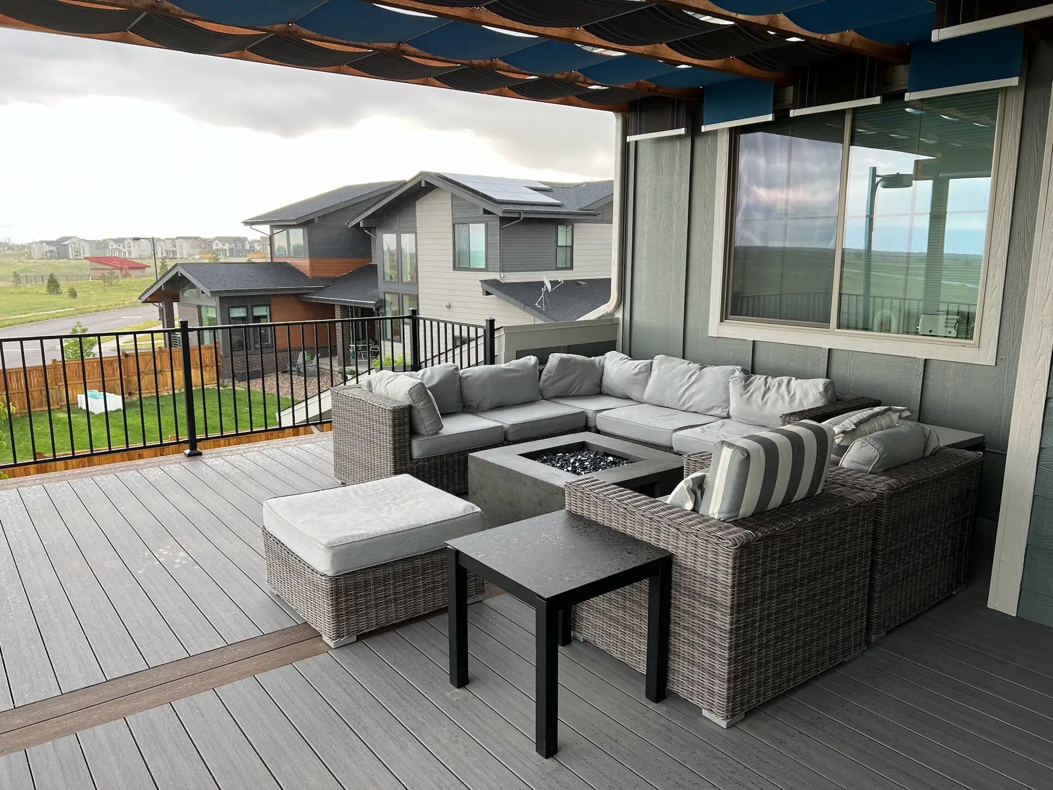 composite decking brands A covered outdoor deck with gray composite flooring features an L-shaped brown wicker sectional with gray cushions, an ottoman, a small black side table, and a square stone fire pit. The deck has a retractable blue-and-black striped awning and a black cable railing.